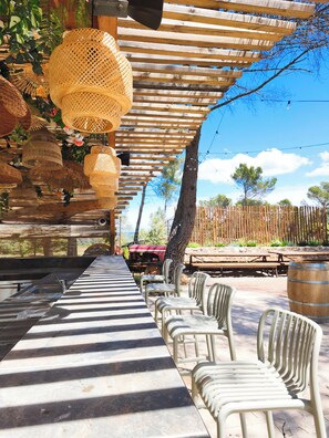Un espacio comedor al aire libre cubierto con una mesa larga, sillas, lámparas de mimbre colgantes y una valla de madera al fondo.