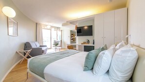 Habitación de hotel moderna con una cama grande, un área de descanso con una silla y una mesita, televisión y una kitchenette.
