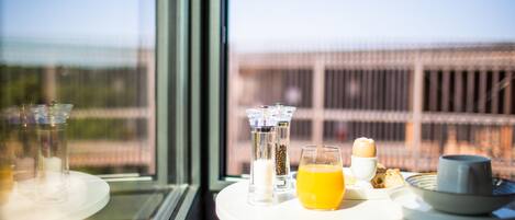 Mesa de café da manhã com copo de suco de laranja, xícara e prato com torradas e uma pequena tigela, em frente a uma janela com vista para um edifício.