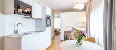 A modern kitchen with white cabinetry, a built-in oven, and a stainless steel sink. There is a dining area with a round table and a vase of flowers.