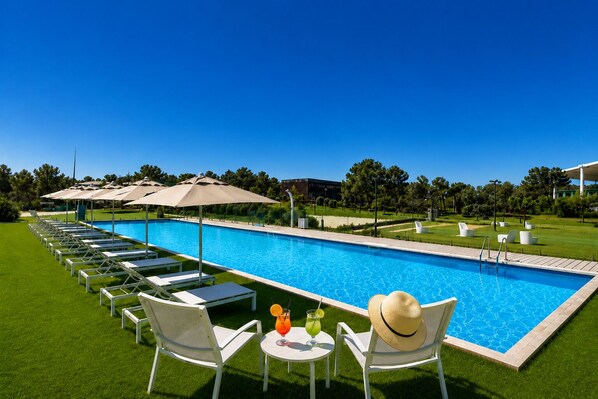 Una piscina con sillas de descanso y sombrillas, rodeada de césped y árboles.