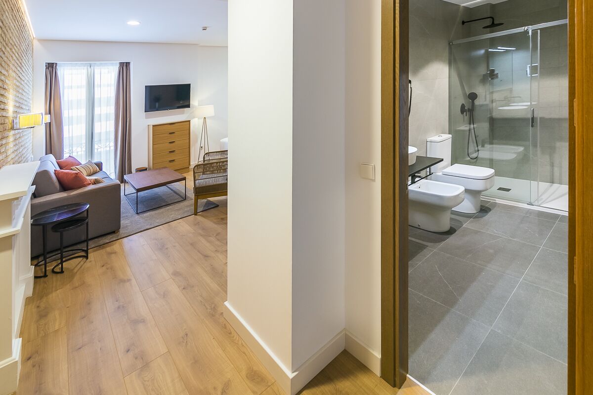 A modern hotel room with a sofa, a TV, a desk, and a glass-enclosed shower area.