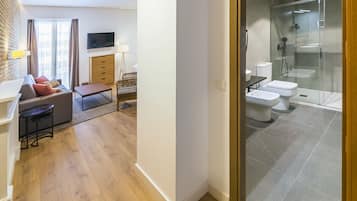 A modern hotel room with a sofa, a TV, a desk, and a glass-enclosed shower area.