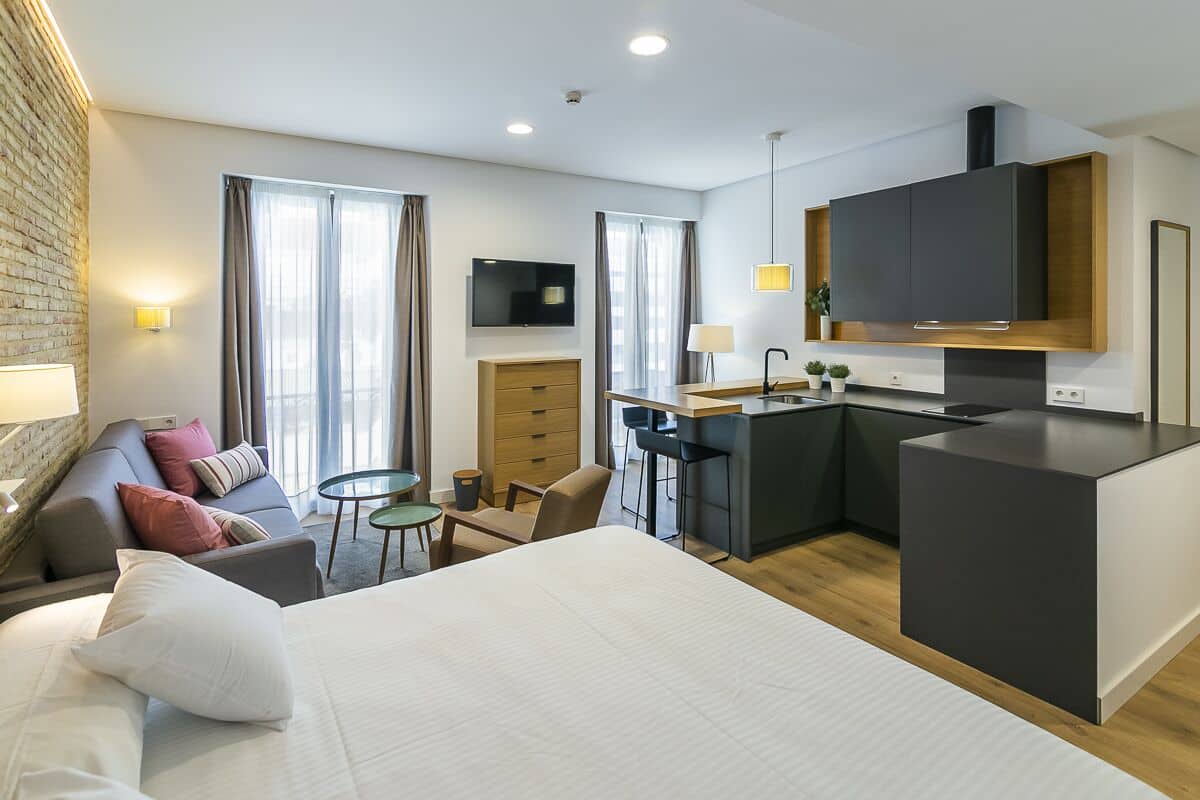 A modern hotel room with a bed, a sofa, a small dining area, a TV, and a kitchenette.