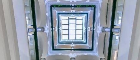 Un interior moderno con un ascensor de vidrio central, suelo de madera e iluminación colgante.