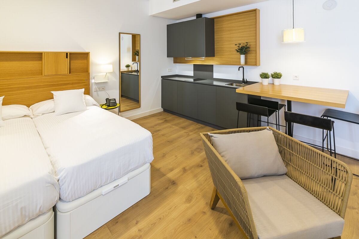 A modern hotel room with a bed, a wicker chair, a small kitchen area, and a dining table.