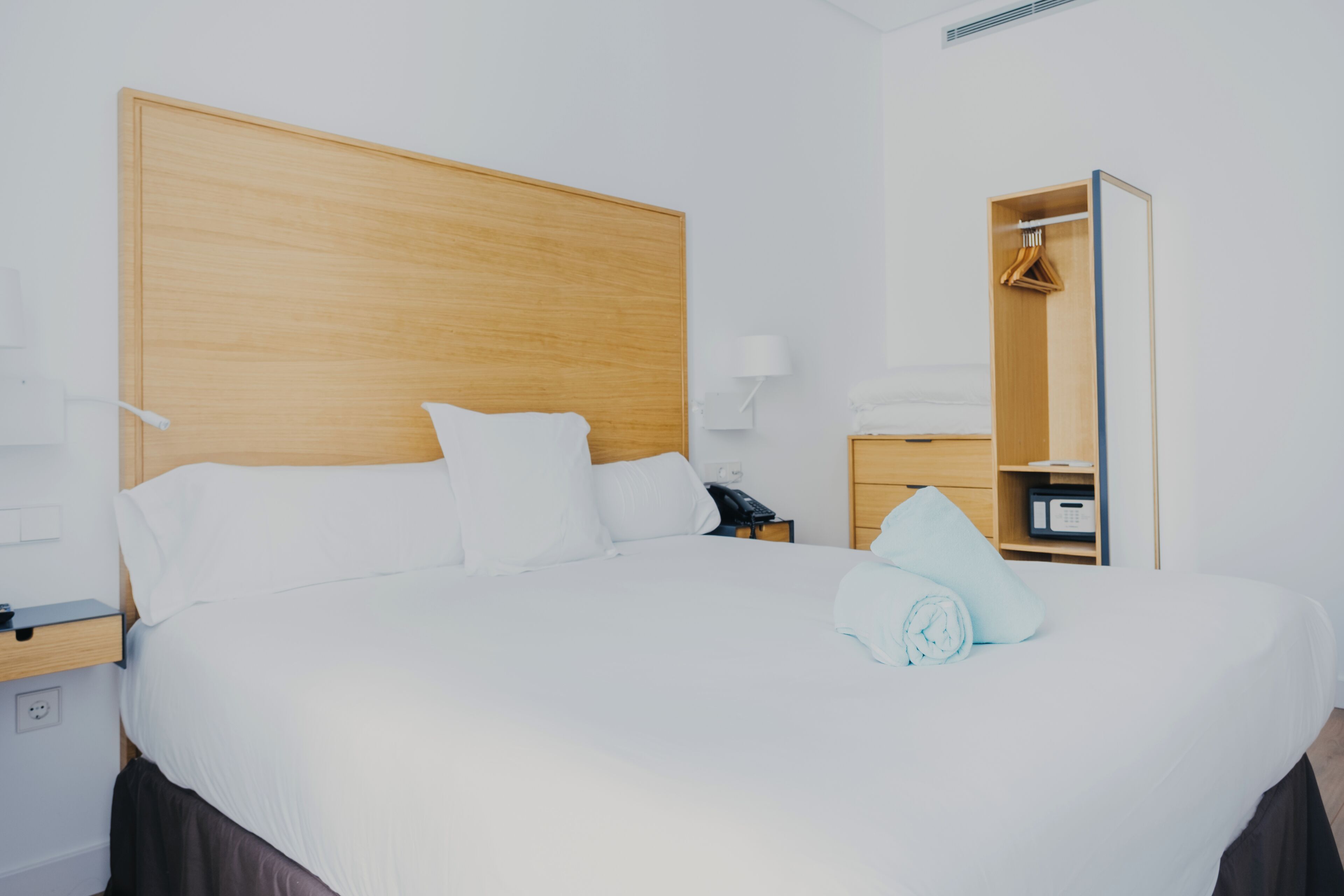 Una cama bien hecha con ropa blanca, un cabecero de madera, una mesita de noche con lámpara y un armario empotrado.
