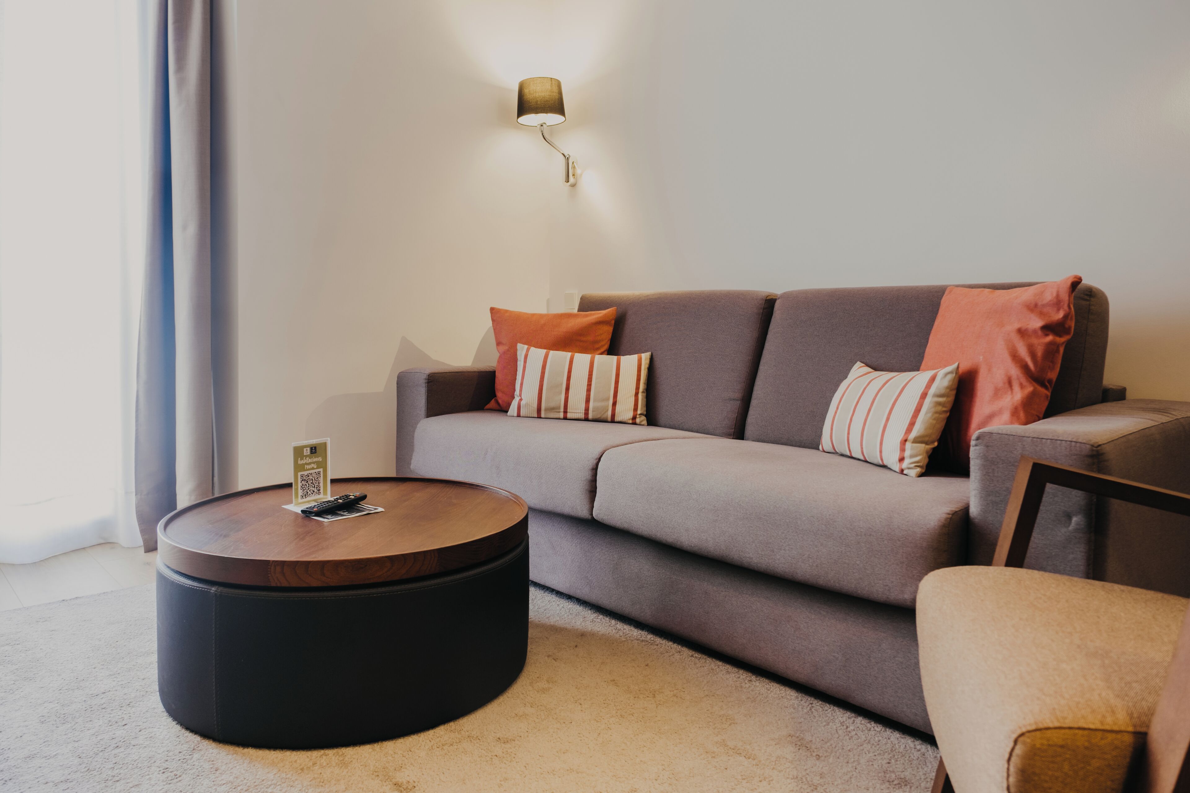 A modern living room with a grey sofa, two striped pillows, a round wooden coffee table, and a wall-mounted lamp.