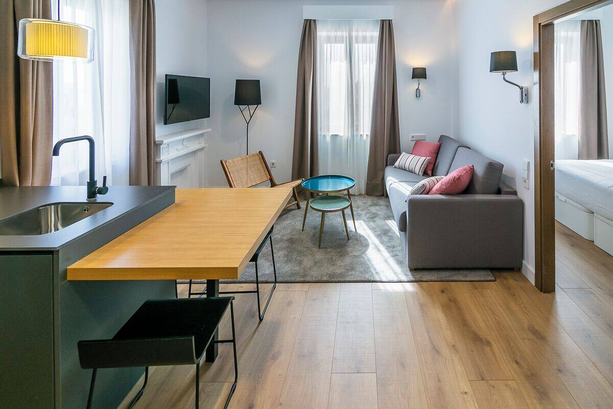 A modern hotel room with a wooden dining table, a sofa, a small blue table, and a TV mounted on the wall.