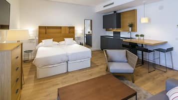 Habitación de hotel moderna con cama doble, sofá, mesita, zona de cocina y televisor.