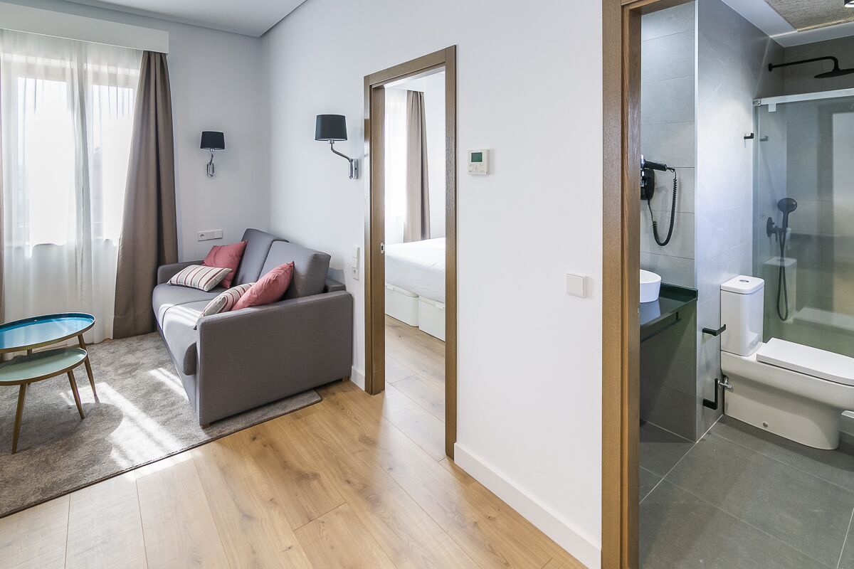 Ein modernes Hotelzimmer mit grauem Sofa, einem kleinen runden Tisch, einer Duschkabine aus Glas sowie einem separaten Bad mit Toilette und Waschbecken.