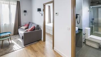 Una habitación de hotel moderna con un sofá gris, una mesita redonda pequeña, un box de ducha de vidrio y un baño separado con inodoro y lavabo.