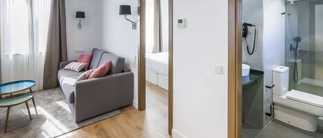 Ein modernes Hotelzimmer mit grauem Sofa, einem kleinen runden Tisch, einer Duschkabine aus Glas sowie einem separaten Bad mit Toilette und Waschbecken.