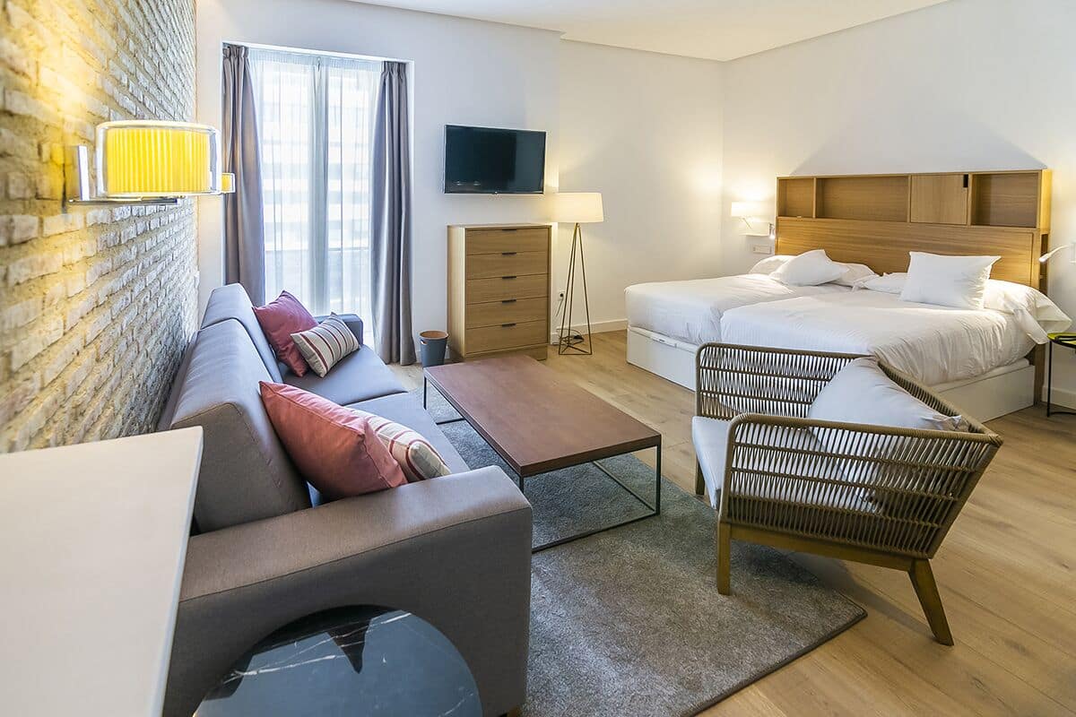 A modern hotel room with a sofa, a bed, a small table, and a chair. There is a TV mounted on the wall and a lamp beside the bed.