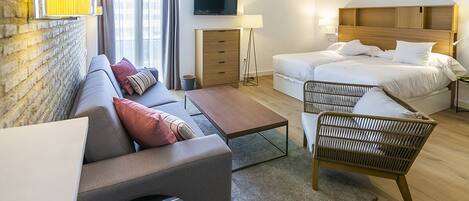 A modern hotel room with a sofa, a bed, a small table, and a chair. There is a TV mounted on the wall and a lamp beside the bed.