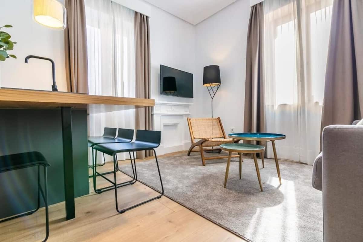 A modern hotel room with a wooden desk, two chairs, a small round table, a flat-screen TV, and a fireplace.