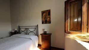 Un dormitorio con cama, mesita de noche, armario pequeño, un cuadro en la pared y ventana con marco de madera.