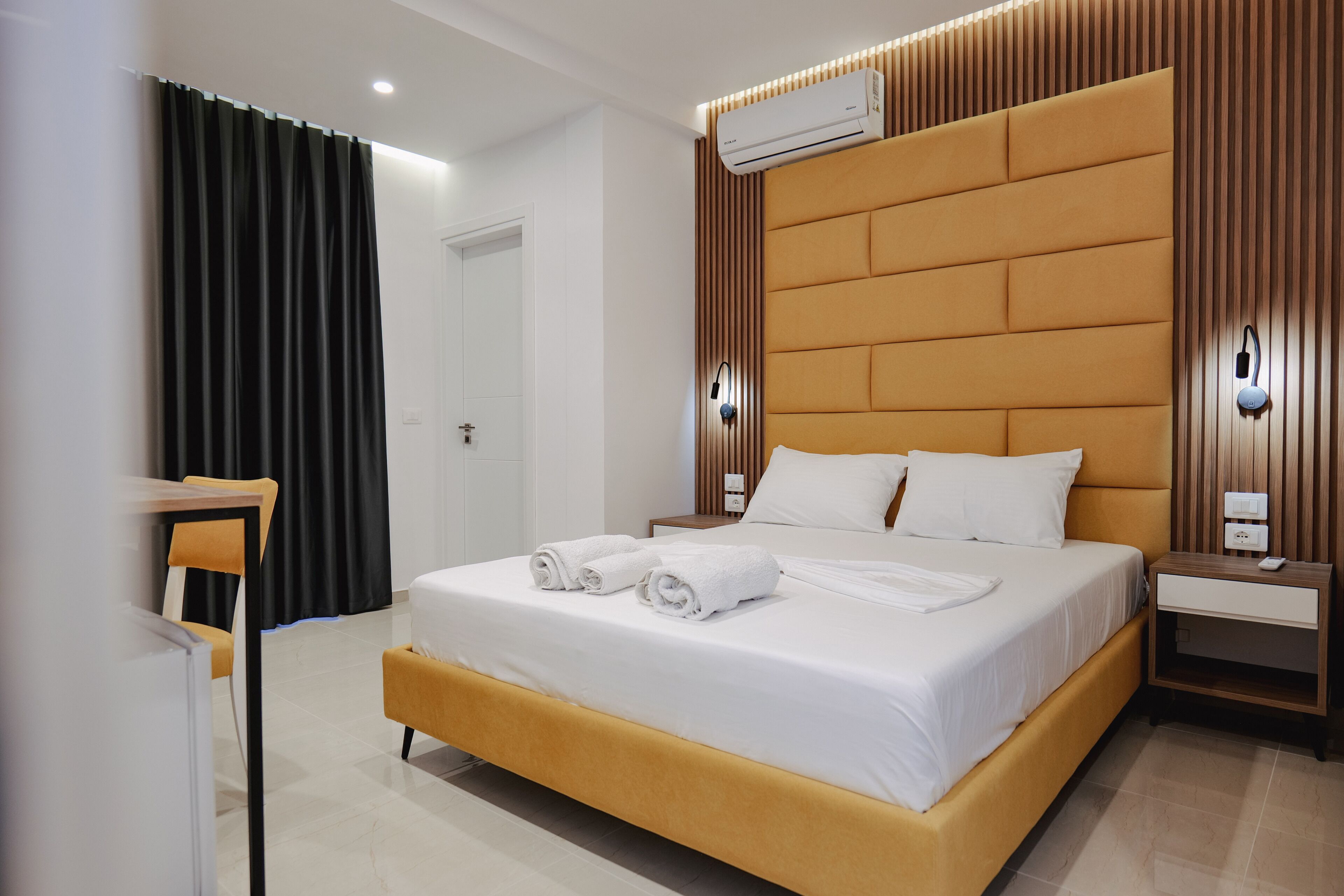 A modern hotel room with a large bed, a bedside table, an air conditioning unit, and a wall-mounted light fixture.