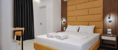 A modern hotel room with a large bed, a bedside table, an air conditioning unit, and a wall-mounted light fixture.