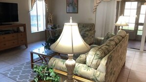 A living room with a sofa, a coffee table with a lamp, a television, and a patterned armchair.