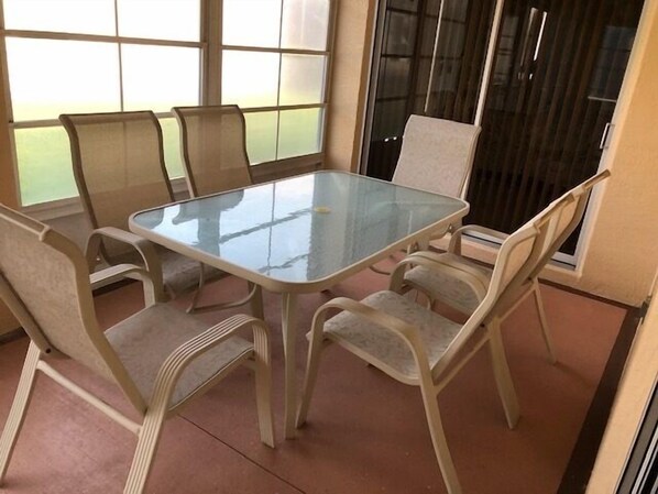 A glass-top table with six chairs in a room with large windows and a view of a green area outside.