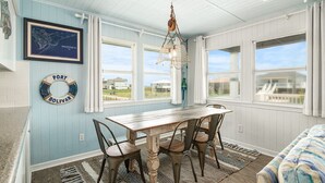 Un espace repas avec une table et des chaises en bois, un luminaire suspendu et une vue sur l’extérieur grâce à de grandes fenêtres.