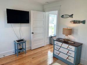 Een kamer met een wandgemonteerde flatscreen-tv, een piano, een houten vloer, een witte deur, een raam met zonwering, een lamp en een dressoir met lades.