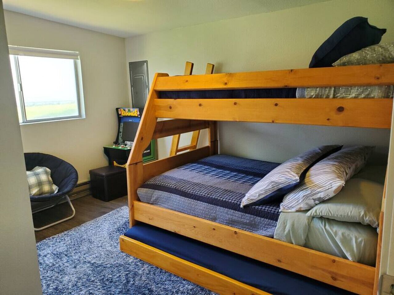 Una cama litera con colcha azul y gris a cuadros, cabecera de madera y ventana con vista al exterior.