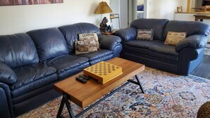 Ein Wohnzimmer mit zwei dunklen Ledersofas, einem Holztisch mit Schachbrett und einem bunten Teppich.