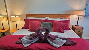 Ein ordentlich bezogenes Bett mit einer herzförmigen Handtuchplatzierung, roter und grauer Bettwäsche sowie zwei Nachttischlampen.