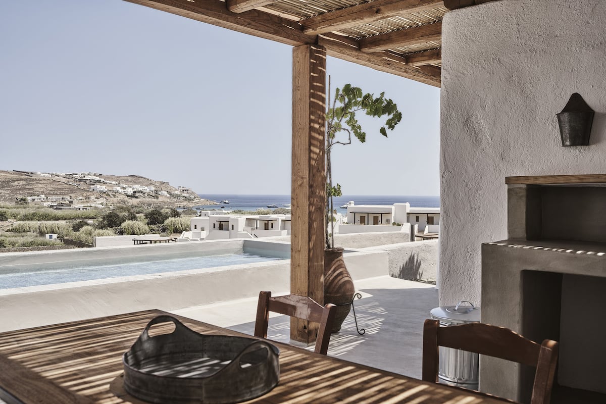 A wooden table with chairs, a potted plant, and a view of a coastal landscape with buildings and a pool.