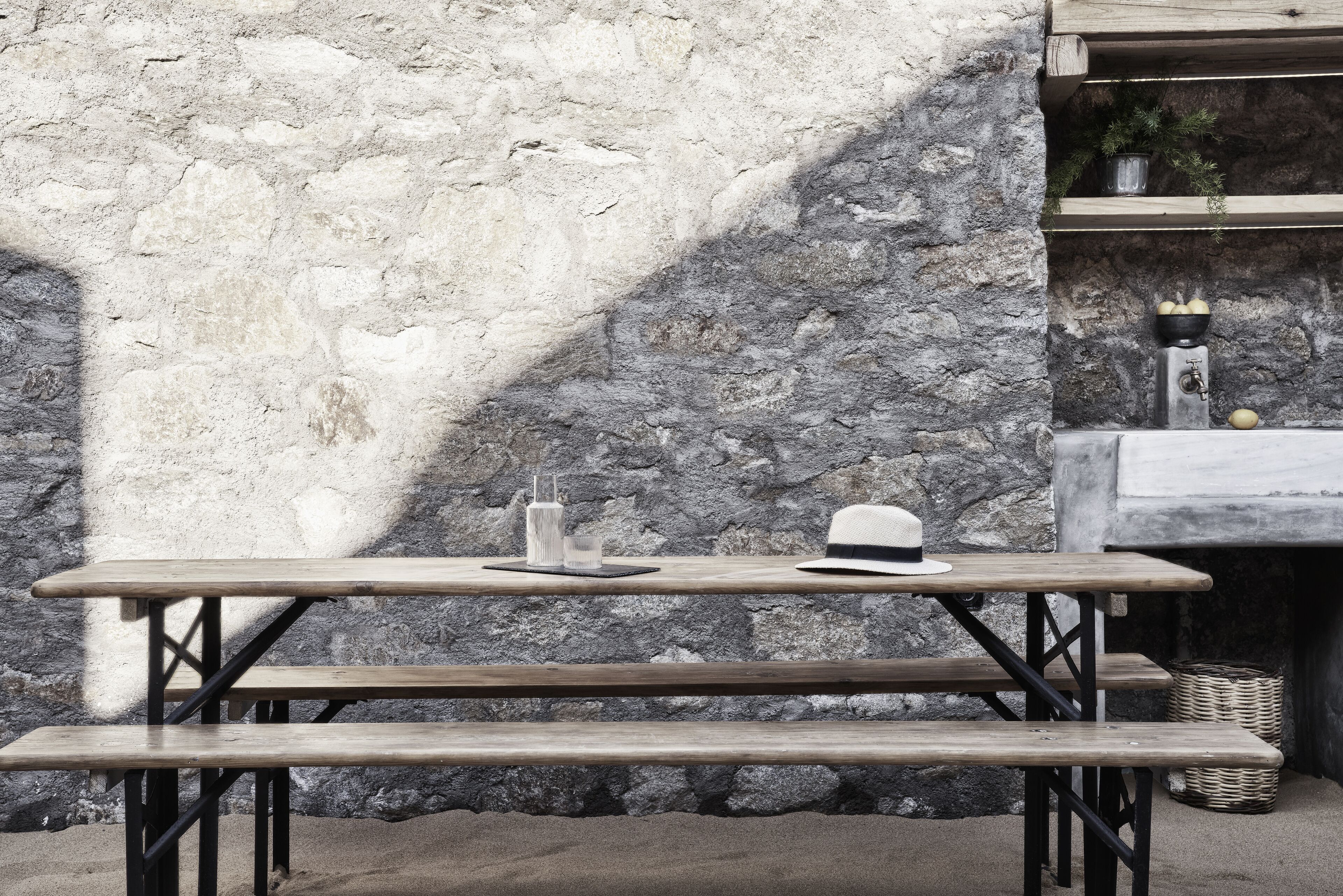 A wooden picnic table with a hat and glasses on it, set against a stone wall.