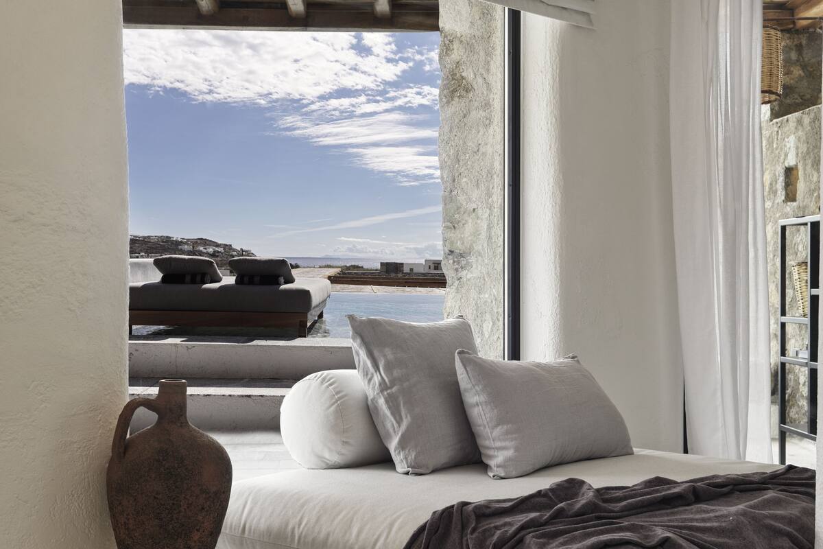 A room with a view of the sea, a white sofa with cushions, a large vase, and a window with curtains.