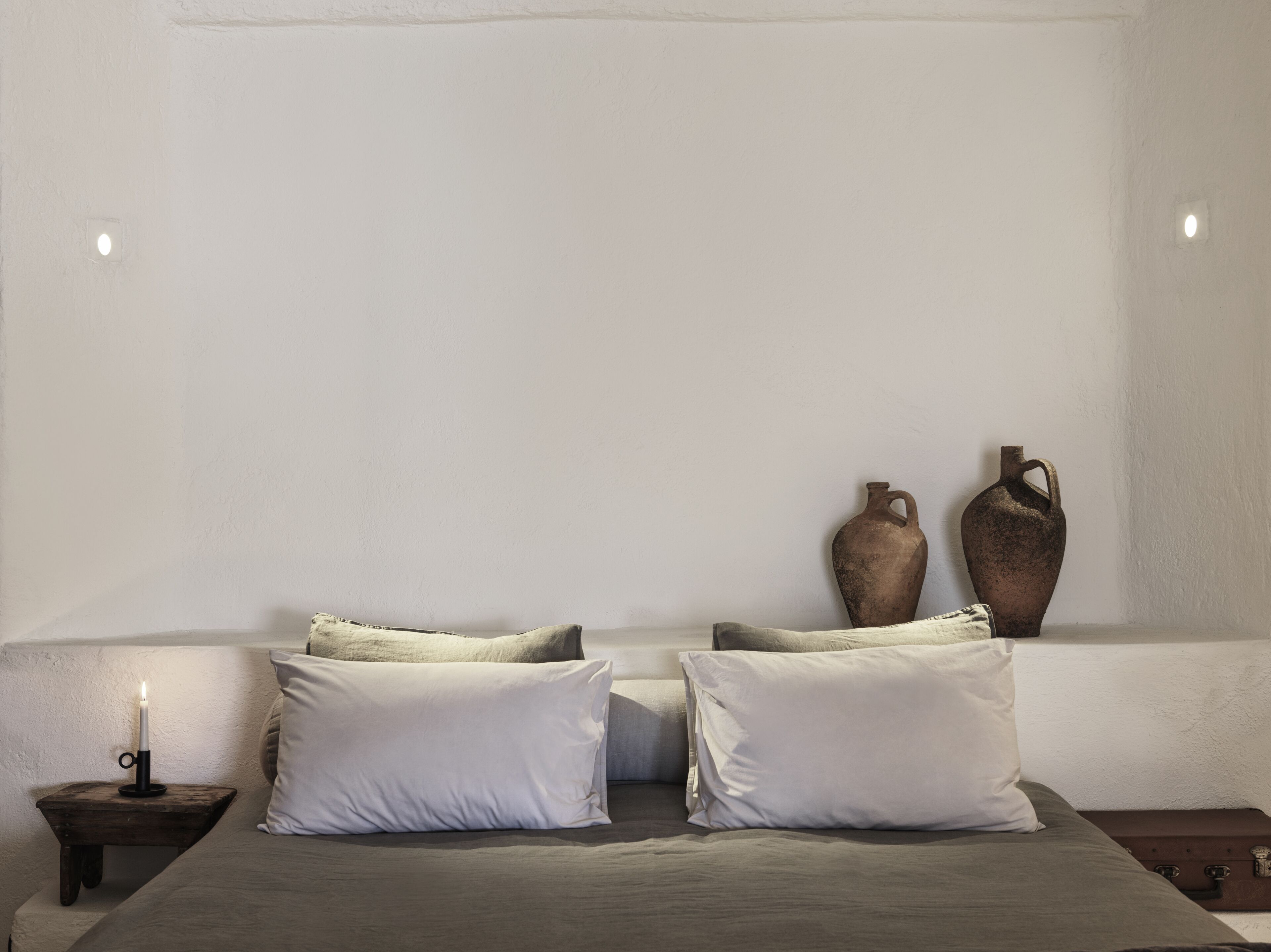 Een slaapkamer met een bed, twee kussens, een nachtkastje met een kaars en twee vazen aan de muur.