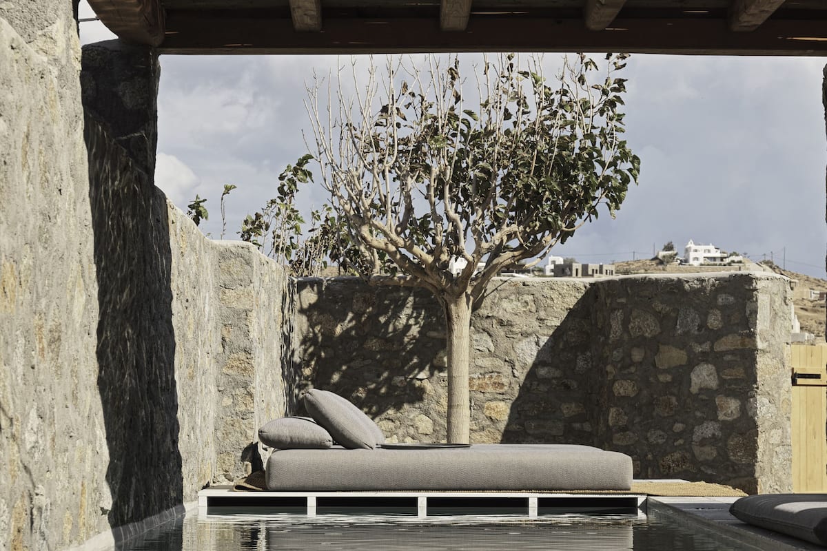 A poolside lounge area with a built-in bench and cushions, a tree, and a stone wall in the background.