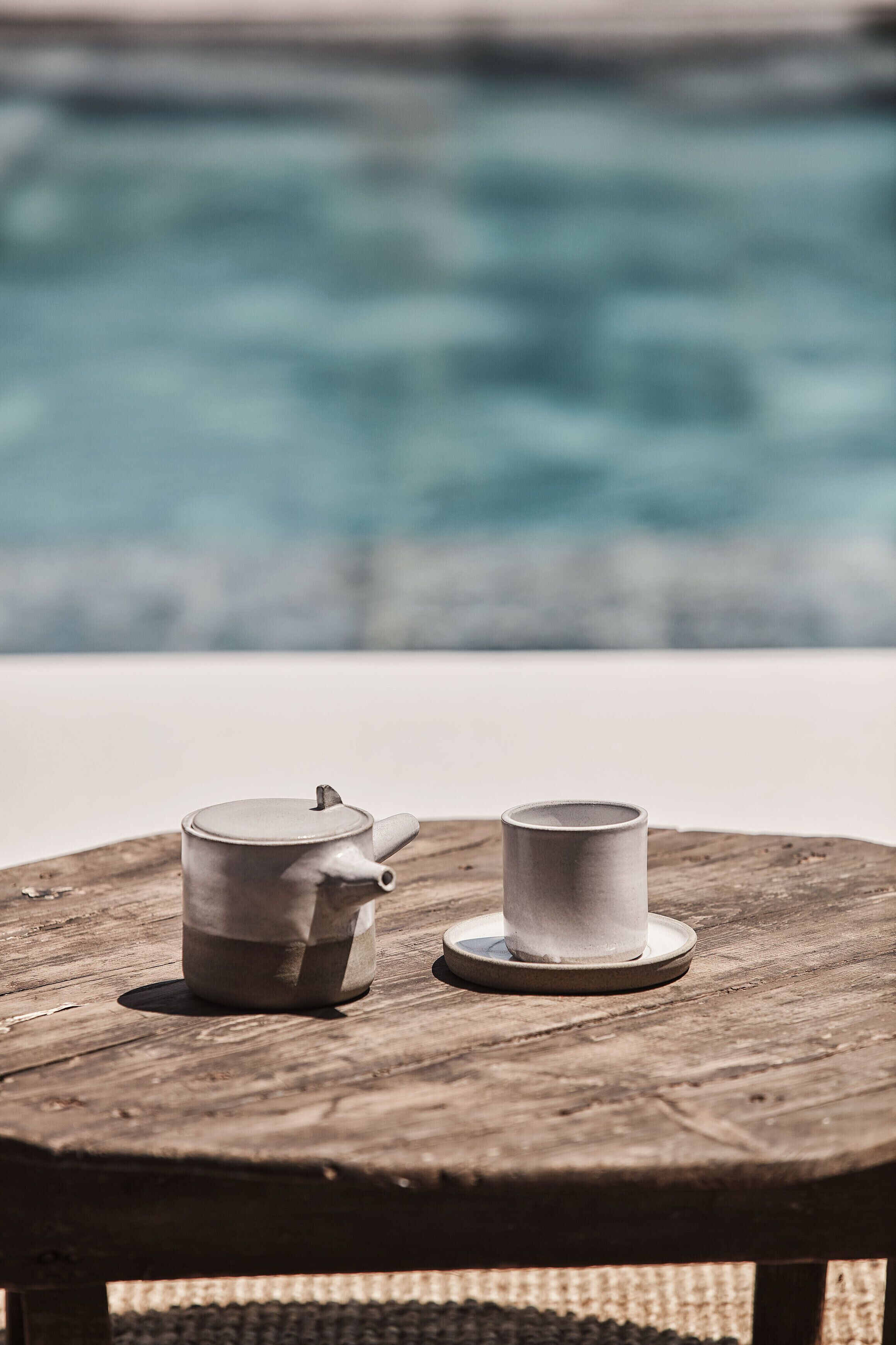 A wooden table with two white ceramic cups and a teapot by a pool.