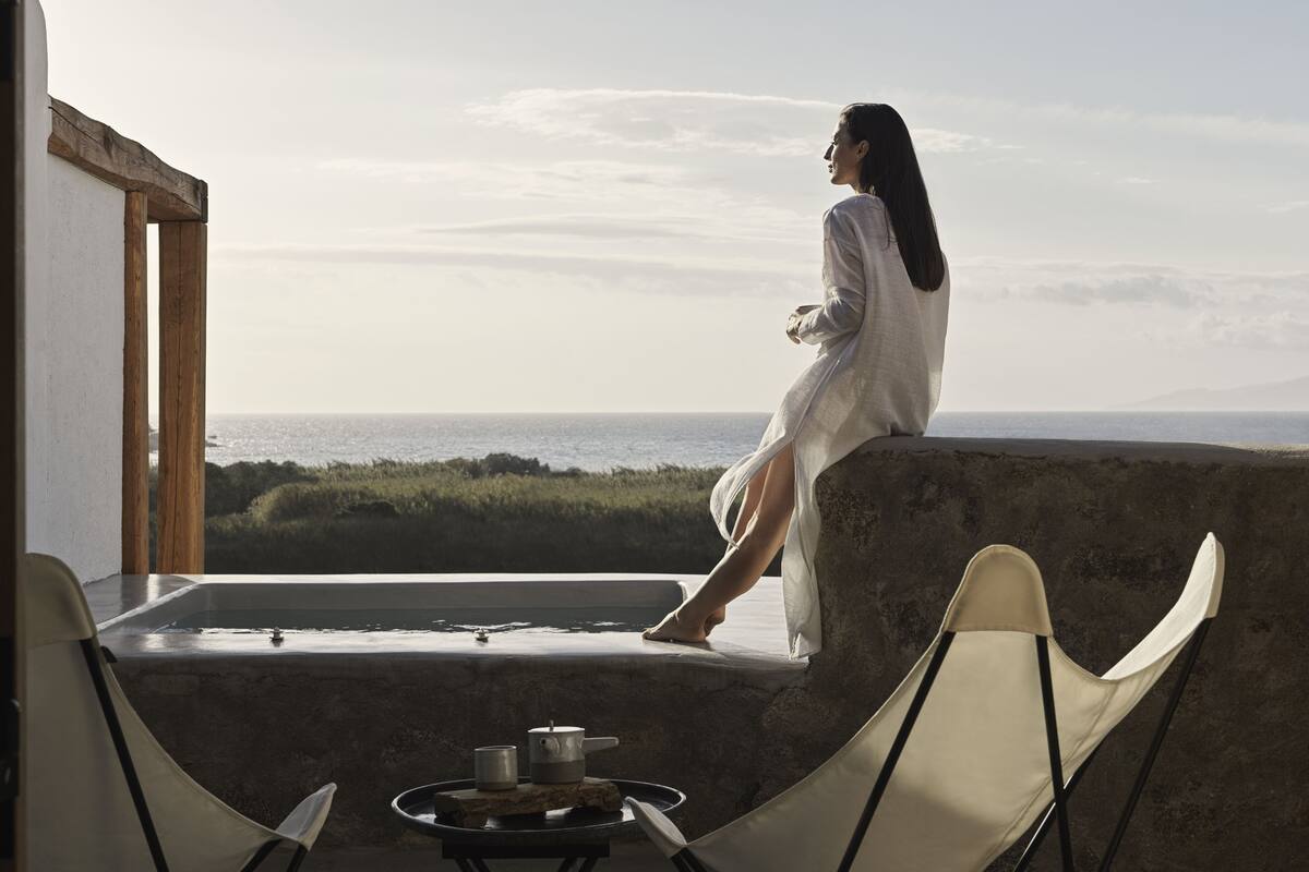 A woman sitting by a window overlooking a beach, with a table and chairs in the foreground.