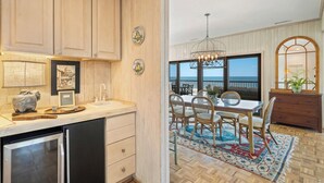A kitchen with wooden cabinets, a sink, and a refrigerator. There is a dining area with a table and chairs, a chandelier, and a view of the ocean. - Penthouse Oceanfront Villa at Inlet Point on Litchfield Beach (Pawleys Island)