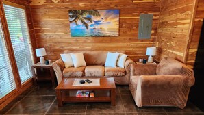 Una habitación acogedora con paredes de madera, un sofá grande, dos mesitas de noche con lámparas y un cuadro con una escena de playa.