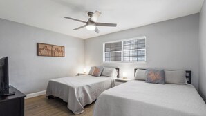 Ein Schlafzimmer mit zwei Betten, einem Deckenventilator, einem Fenster mit Jalousien, einem Fernseher und einem Wandbild.