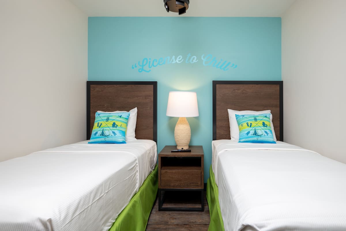 A hotel room with two beds, a wall-mounted lamp, and a wall with a light blue background and a "License to Grill" sign.