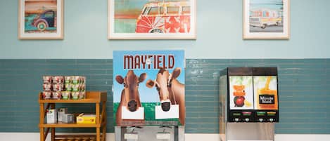 En disk med en dispenser til mælk og juice, en hylde med mælkekartoner, og indrammet kunst på væggen.