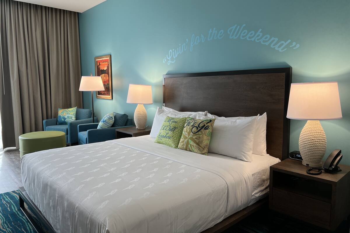 A hotel room with a large bed, two bedside tables with lamps, a blue armchair, a green ottoman, and a wall with a 'Like for the Weekend' sign.