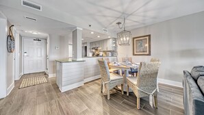 A modern kitchen with white cabinetry, a dining area with wicker chairs, and a grey sofa in a bright, neutral-toned living space.