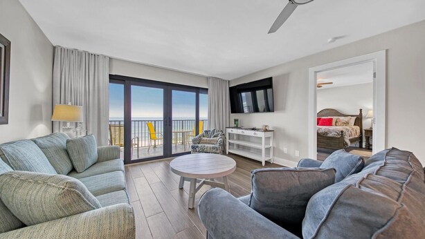 A modern living room with a sofa, armchairs, a small round table, and a TV. A balcony with a view of the sea is visible through the glass doors.