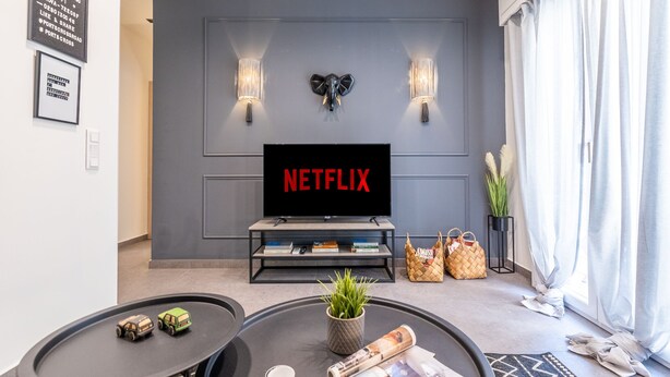 A modern living room with a flat-screen TV mounted on a grey wall, a coffee table with a plant, and a bookshelf with books and decorative items. - Ports Crossroad E (Πειραιάς)