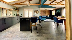 Una cocina moderna con una isla central, un comedor con sillas azules y una sala de estar con un sofá y un reloj de pared.