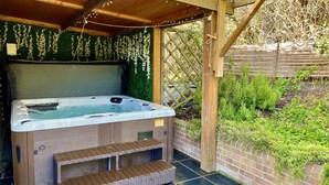 Un jacuzzi bajo una pérgola de madera con un muro verde de fondo y un muro de ladrillo con plantas.