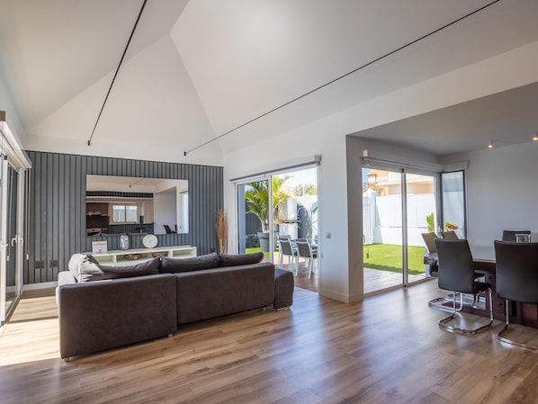 Un salon moderne avec un canapé d’angle gris, un plancher en bois et une vue sur une cuisine et une terrasse extérieure.
