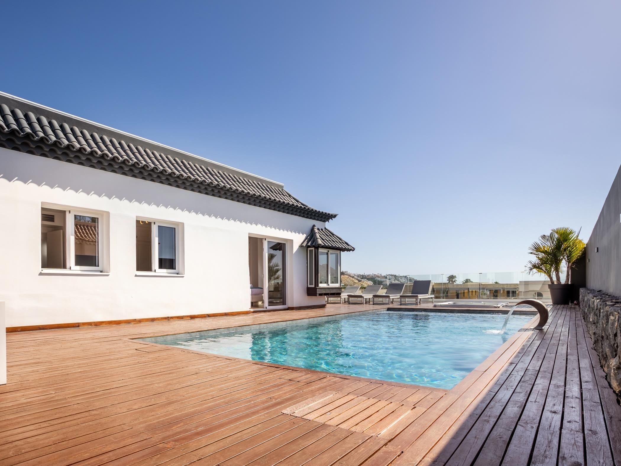 Un espace aménagé au bord de la piscine, avec une terrasse en bois, une piscine et un bâtiment doté d’un toit en tuiles et de murs blancs.